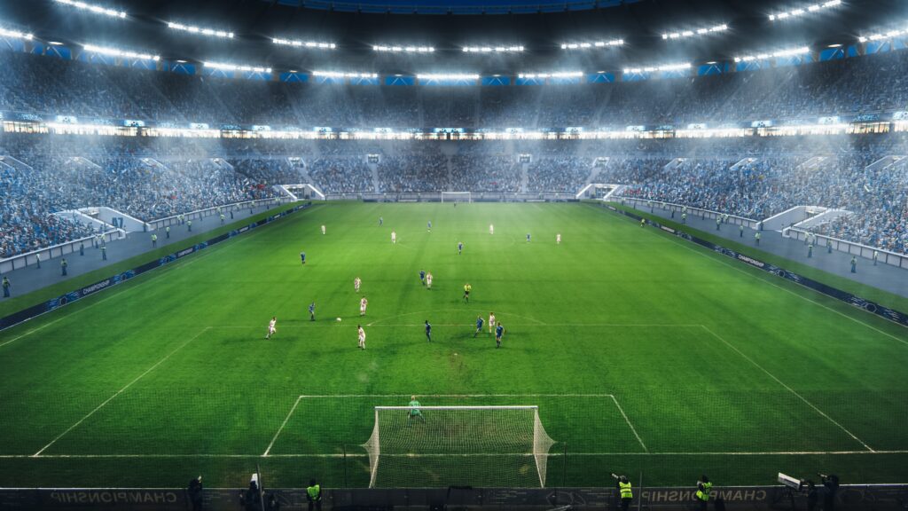 View of a football stadium from the goalpost, full to capacity and with players on the pitch.